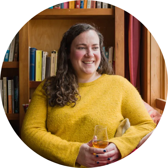 Portrait of a smiling Nora Ganley-Roper wearing a yellow sweater, sitting in front of a bookcase, with a glass of whiskey in-hand.